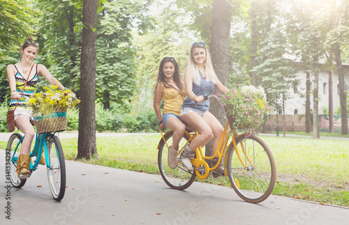 Happy boho chic girls ride together on bicycles in park