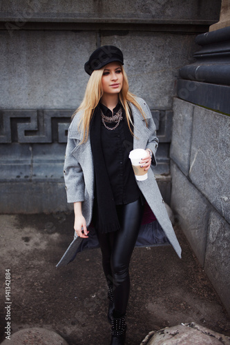 Young, hip and attractive blonde walking around the city with coffee to go, girl in a stylish hat and a gray coat