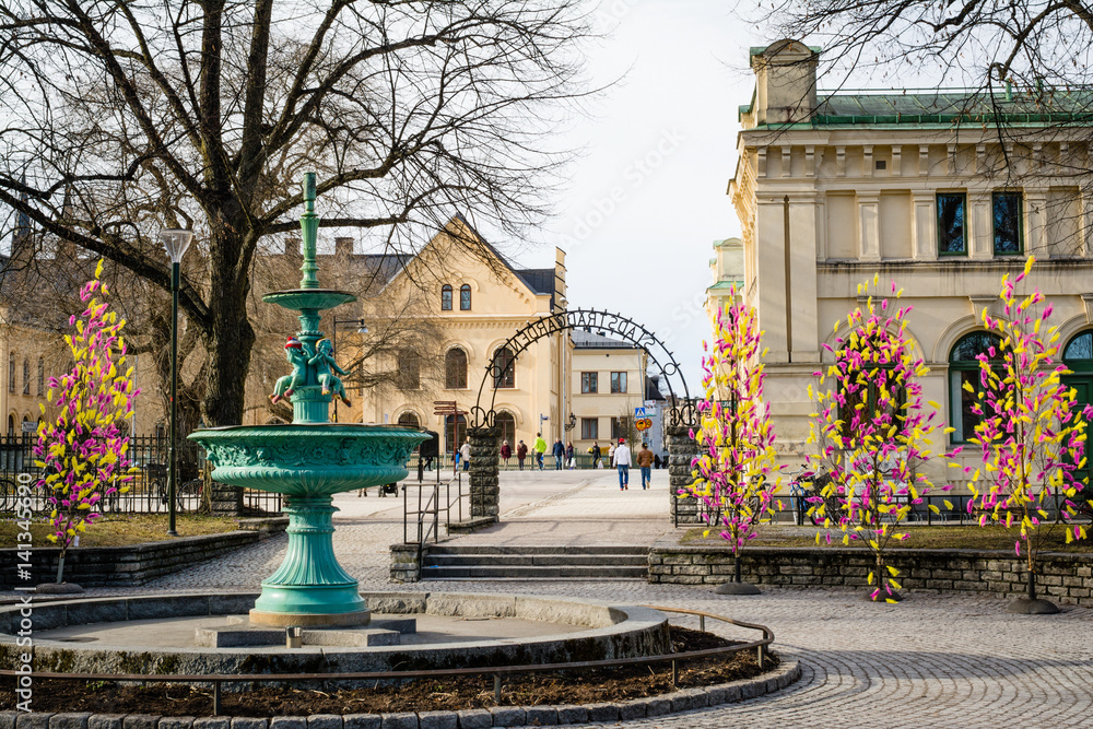 Street view of inactive fountain with traditional colourful feathers on trees for Easter decorations in Uppsala, Sweden, Europe