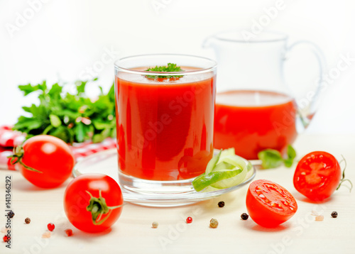 tomato juice in glass, jug with greenery, basil, cutted tomato fruit and dry pepper on light table, concept vegetarian food, close up
