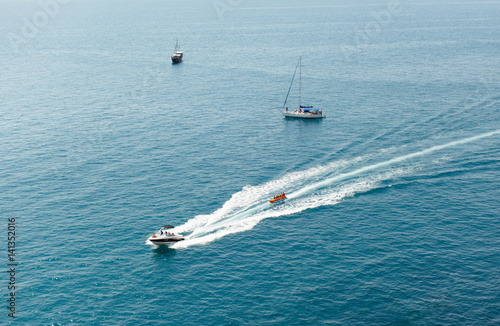 The motor boat sails on the black sea and carries on people's inflatable banana in life jackets. In the background are two sailing yachts. Horizontal shot, summer, rest, daylight.