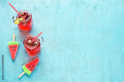 Two portions of homemade watermelon lemonade in mason jar with red striped straw and watermelon popsicles on a blue background