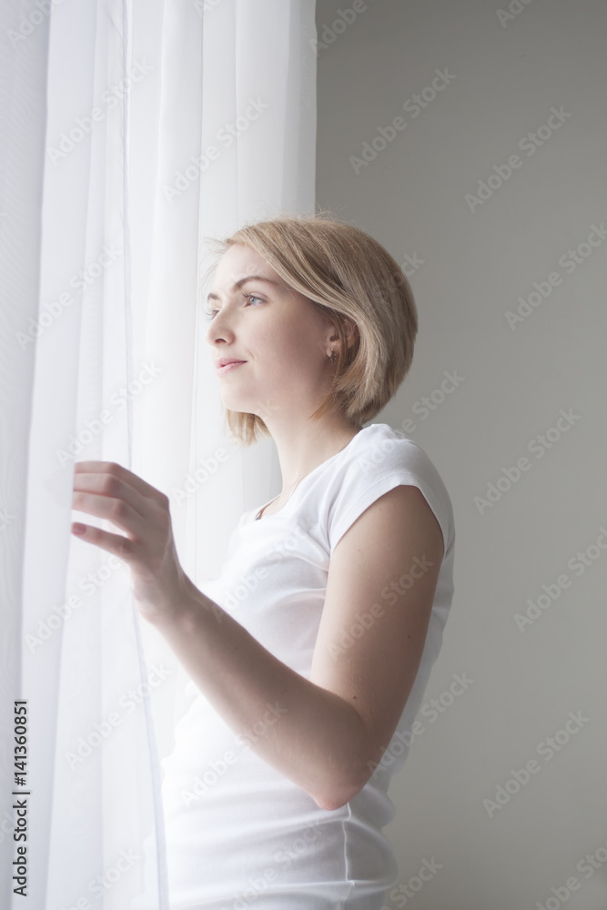 Young pretty smiling girl opening curtains in the morning to welcome spring Young pretty smiling girl opening curtains in the morning to welcome spring