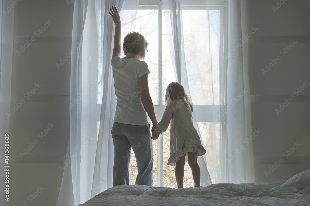 Woman and little girl, wake up and open the curtains in the morning, back view Woman and little girl, wake up and open the curtains in the morning, back view