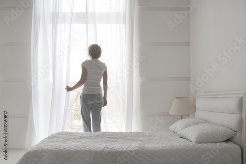Quadro su tela Young pretty girl opening curtains in the morning to get fresh air, back view