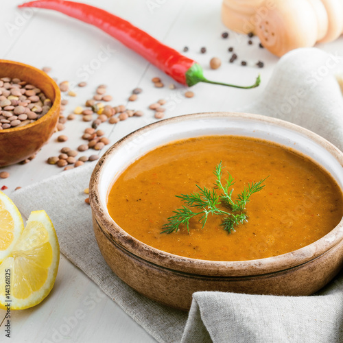 Healthy vegetarian lentil soup in a rustic clay bowl , piece of bread and lemon slices on a white wooden table. Delicious healthy meal served on a canvas tablecloth.