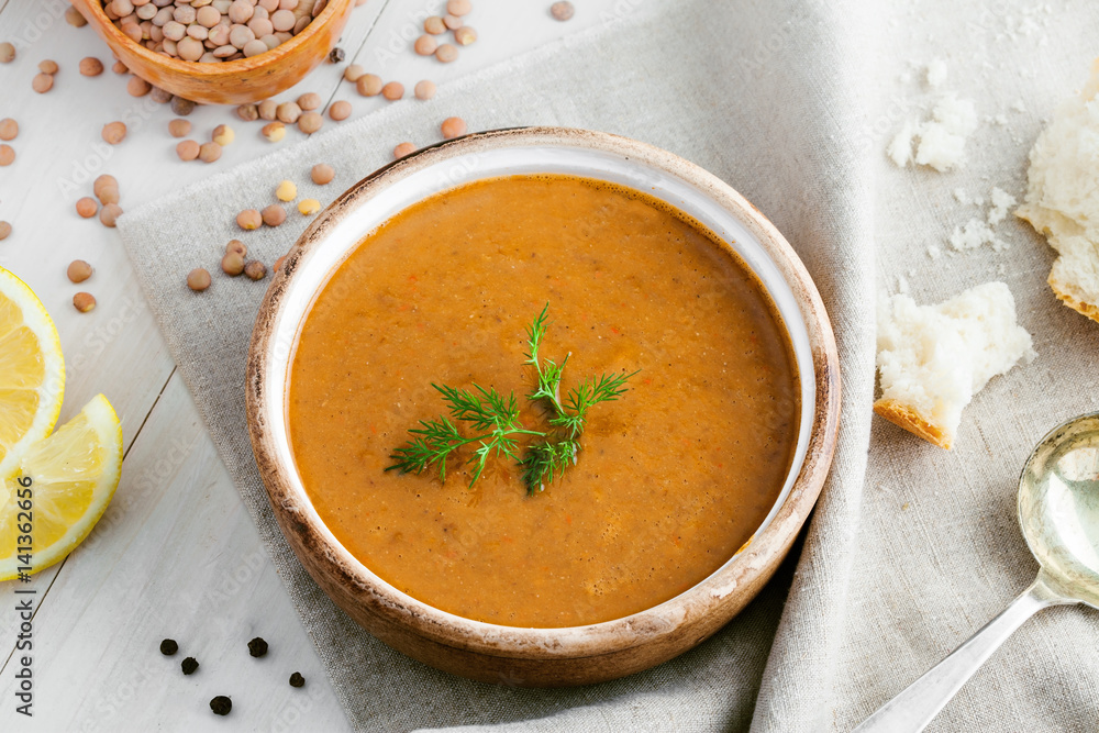 Healthy vegetarian lentil soup in a rustic clay bowl with bread and lemon slices on a white wooden table. Delicious healthy meal served on a canvas tablecloth. Top view shot.