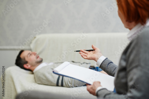 Mature psychologist with clipboard looking at her middle-aged patient and giving him advice how to save his marriage