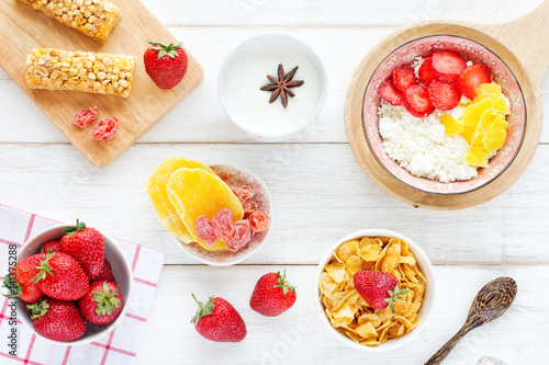 Cottage cheese with berries and dried mango, strawberry on a linen napkin, muesli bars, cornflakes, dried fruits, cup of milk with anise star on a white wooden background.