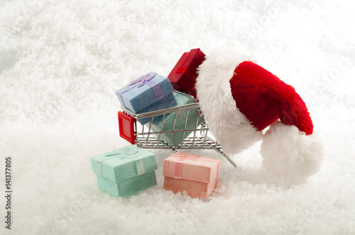 Holiday shopping and Christmas gift exchange concept with shopping cart wearing santa claus hat filled and surrounded by gifts and presents isolated on white snow