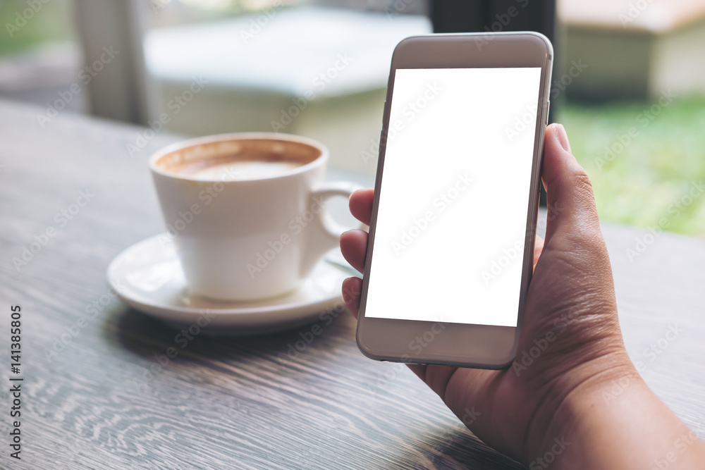 Mockup image of hands holding white mobile phone with blank white screen and hot coffee cup in loft cafe