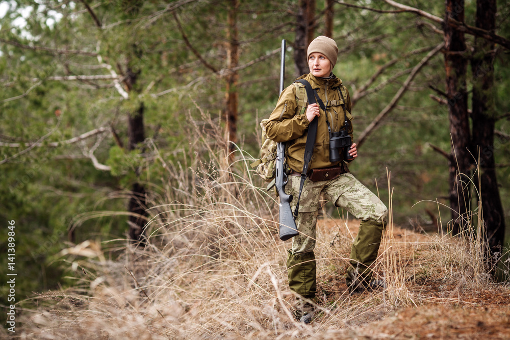 Female hunter in camouflage clothes ready to hunt, holding gun and walking in forest.