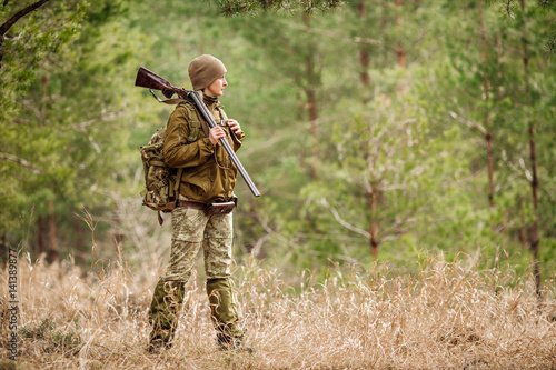 Female hunter in camouflage clothes ready to hunt, holding gun and walking in forest.