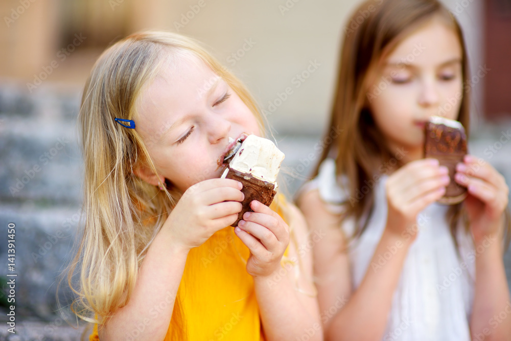 Two cute little sisters eating ice-cream while sitting on the stairs on summer day Two cute little sisters eating ice-cream while sitting on the stairs on summer day