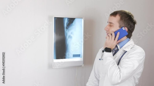 A young male doctor gives a consultation by phone. White wall and a Negatoscope with ribs and rib cage in the background photo