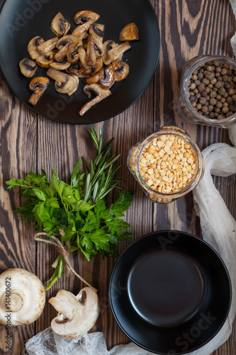 Fried mushrooms in black plate. Dried pea, pepper in glass jars. Fresh parsley and rosemary. Dark wooden table. Toned.