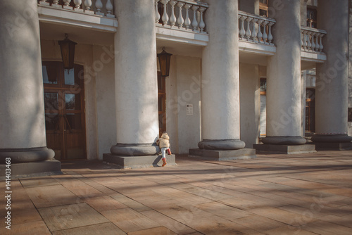 child in a beige jacket, rubber boots and hat in theater columns in the early spring on a sunny day walks
