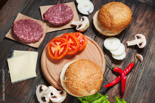 Homemade hamburger ingredients. Raw minced beef, fresh bun, slice of cheese, tomato, onion rings, lettuce on wood background
