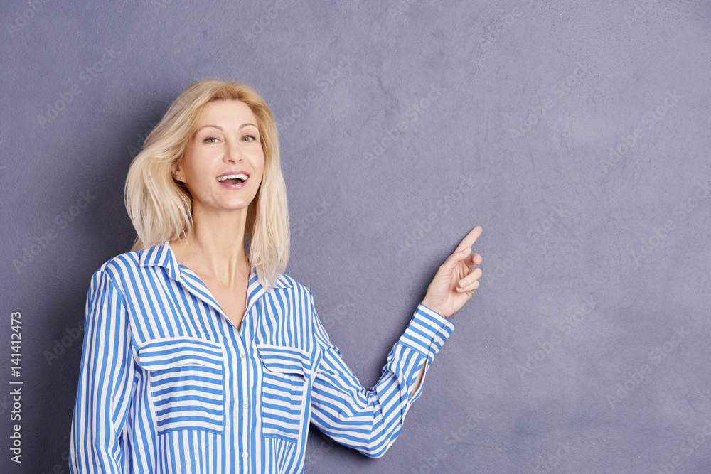 Pointing at copy space. Close-up portrait of a happy business woman pointing at something interesting against at grey wall. Pointing at copy space. Close-up portrait of a happy business woman pointing at something interesting against at grey wall.