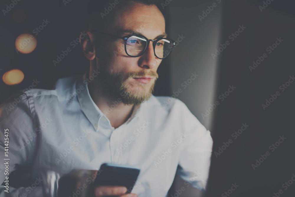 Closeup view of bearded young businessman working on modern loft office at night.Man using laptop and smartphope, blurred background.Horizontal, bokeh effect. Closeup view of bearded young businessman working on modern loft office at night.Man using laptop and smartphope, blurred background.Horizontal, bokeh effect.