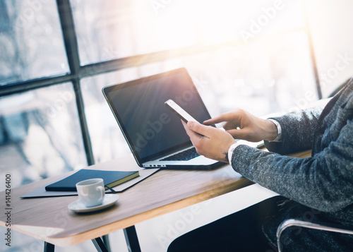 Businessman working at the wood table with laptop at sunny office.Man holding smartphone at hand and texting message.Horizontal, visual effects.Blurred background.