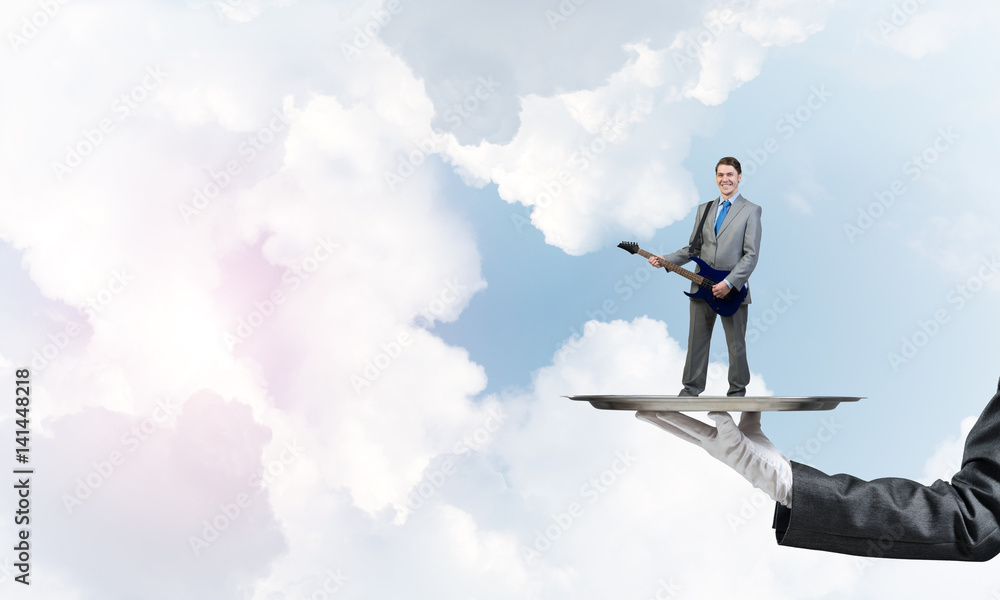 Businessman on metal tray playing electric guitar against blue sky background Businessman on metal tray playing electric guitar against blue sky background