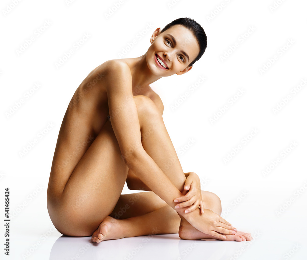 Fototapeta premium Smiling naked woman with perfect body on white background. Beautiful female sitting on the floor touch leg. Beauty & Skin care concept