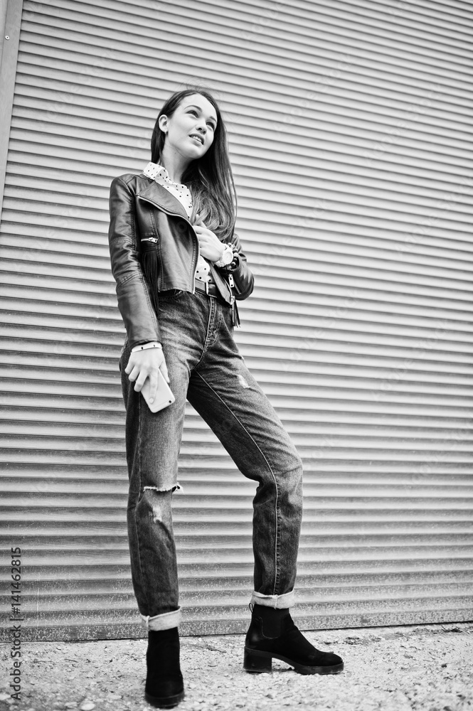 Portrait of stylish young girl wear on leather jacket and ripped jeans with mobile phone at hand background shutter texture. Street fashion model style. Black and white photo.
