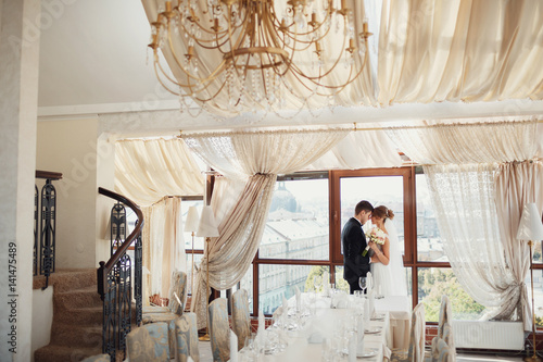 Bride and groom lean to each other tender standing before panoramic window in cosy hotel room