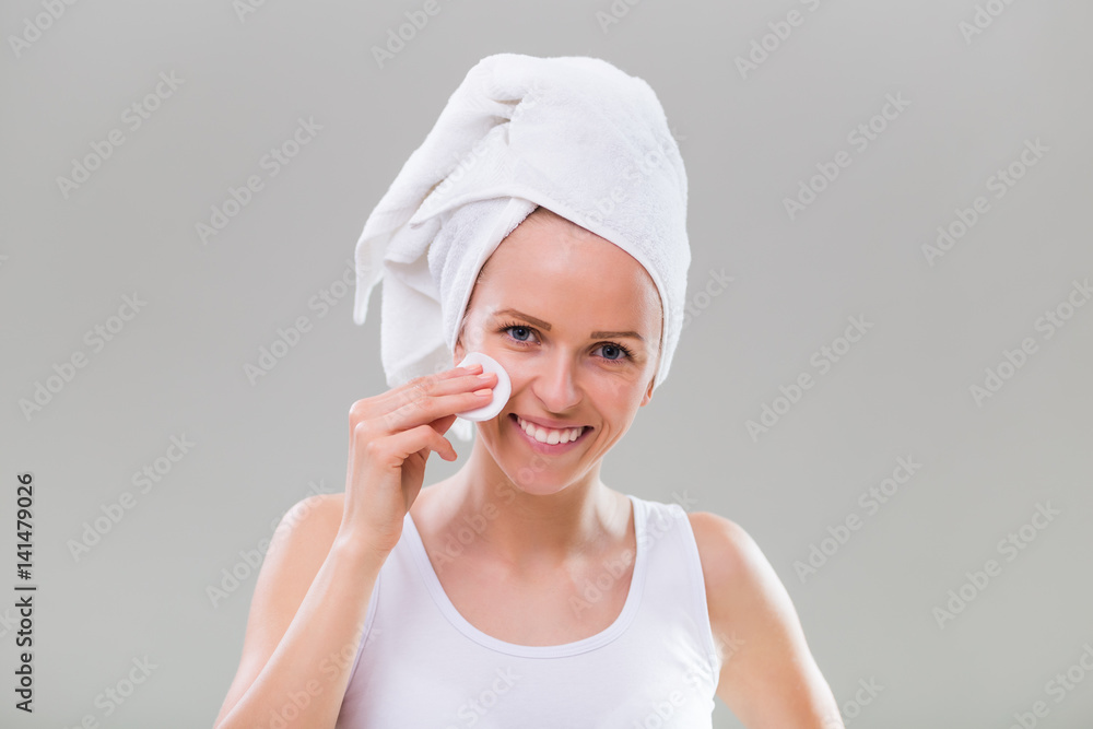 Beautiful young woman cleaning her face with cotton pad on gray background. Beautiful young woman cleaning her face with cotton pad on gray background.