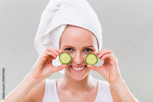 Beautiful young woman showing slices of cucumber on gray background.