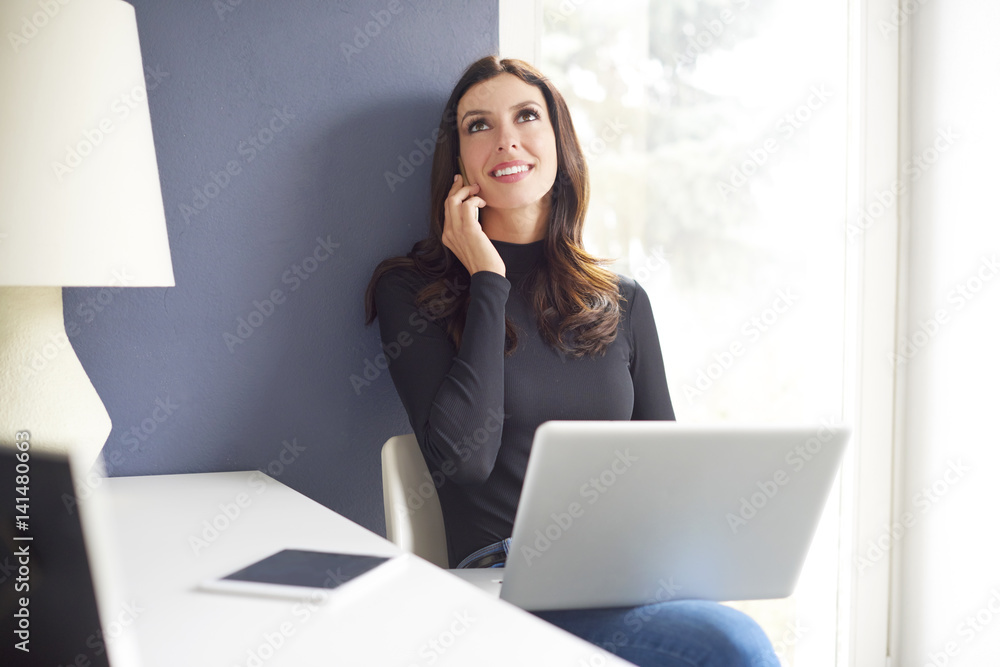 Home office is a great solution. Shot of a happy young woman talking on mobile phone and using laptop while working at office. Home office is a great solution. Shot of a happy young woman talking on mobile phone and using laptop while working at office.