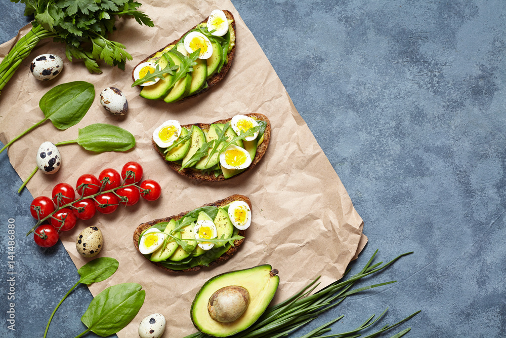Sandwiches with avocado, spinach, guacamole, arugula, tomato and quail eggs on parchment on a concrete background. Spring food. Healthy breakfast, lunch. Low carb diet. Flat lay. Copy space.