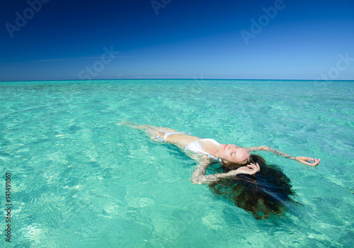 Beautiful young brunette woman with long hair dressed in white swimsuit lies on the crystal azure water of caribbean sea