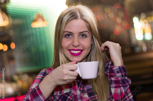 Beautiful young woman holding a cup of coffee looking at camera smiling. Cheerful female enjoying a cup coffee at cafe.