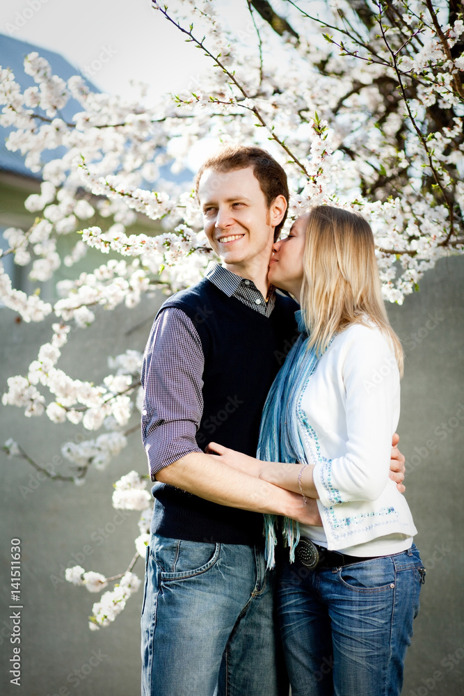 Tender couple in love under blooming cherry tree with white flowers in the park in spring. Happy girl and guy smile and hug outdoor, they are wearing in jeans, blue shirt, white jacket. Tender couple in love under blooming cherry tree with white flowers in the park in spring. Happy girl and guy smile and hug outdoor, they are wearing in jeans, blue shirt, white jacket.