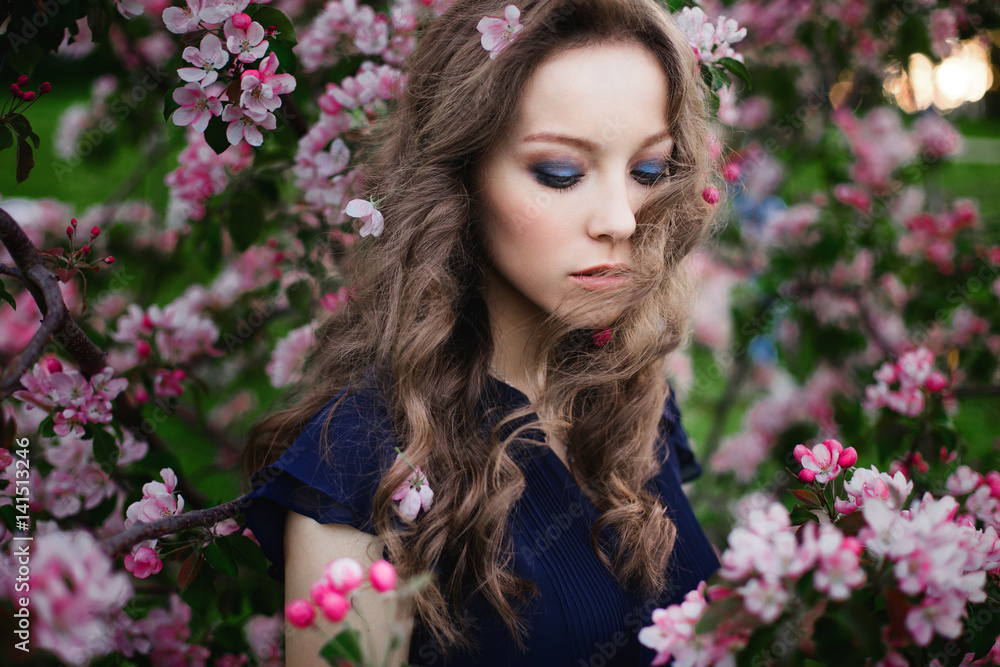 Portrait of a young curly-haired girl with closed eyes in a blue dress standing among a blossoming apple-tree