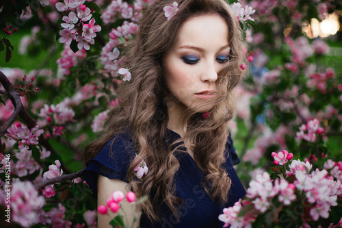 Portrait of a young curly-haired girl with closed eyes in a blue dress standing among a blossoming apple-tree