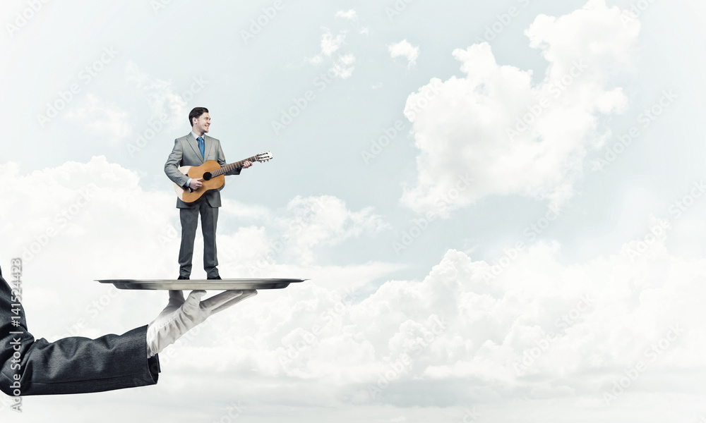 Businessman on metal tray playing acoustic guitar against blue sky background Businessman on metal tray playing acoustic guitar against blue sky background