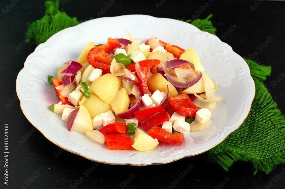 Salad with potatoes, red pepper, onion, basil and feta cheese on white plate on grey cloth on black background