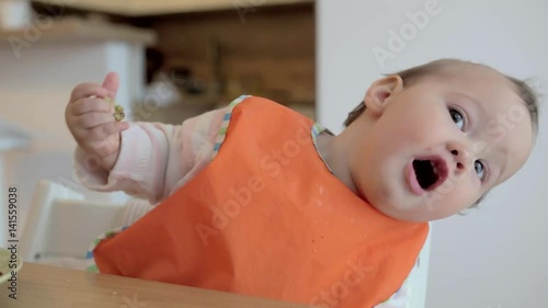 Eleven months old baby feeding herself with her fingers and posing for the camera, moving around, trying to look at the cameraman, baby led weaning; concept of motherhood and parenting