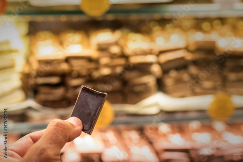 A rectangle of dark chocolate held between fingers against a backdrop of chocolate display