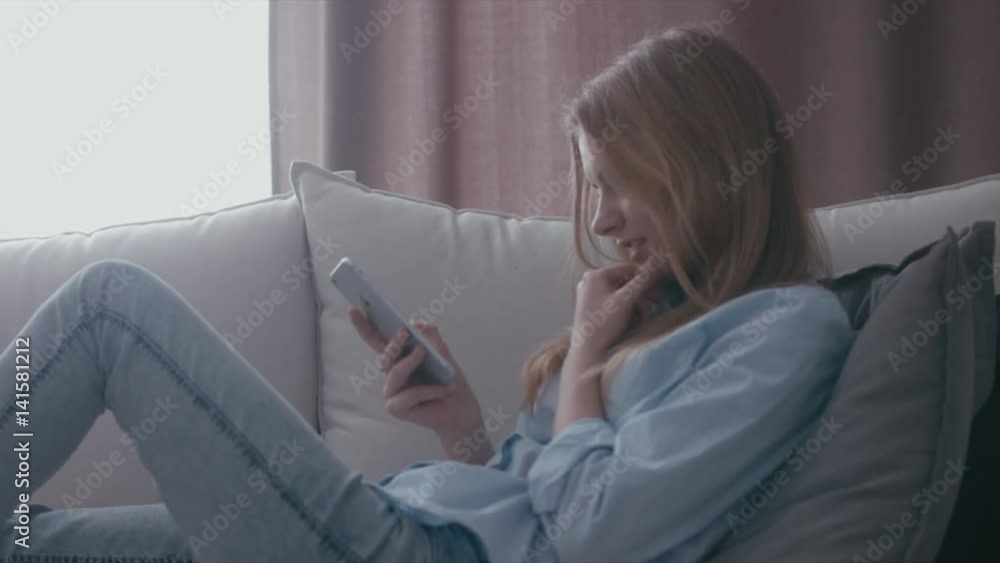 Attractive blonde Caucasian teen girl sitting on a sofa at home and texting on her phone. Smiling, laughing female. Love, friendship concept. 4K UHD RAW edited footage Attractive blonde Caucasian teen girl sitting on a sofa at home and texting on her phone. Smiling, laughing female. Love, friendship concept. 4K UHD RAW edited footage