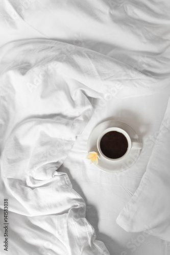A mug of black coffee is standing on a white bed