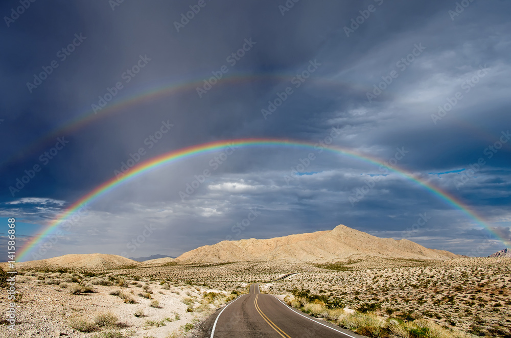 Double rainbow over a desert highway that winds away toward distant hills