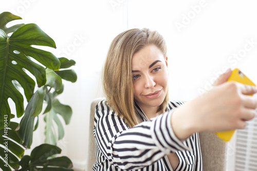 Young blonde makes selfie on her cell phone in the apartment, light interior