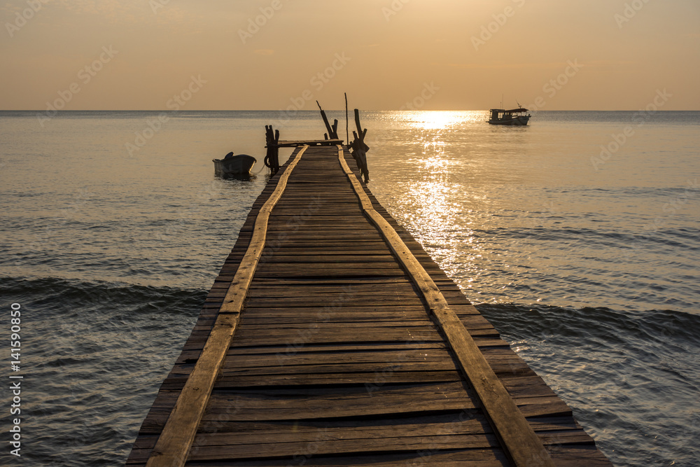 A wonky wooden jetty pier goes out in to a calm sea with small boats around with golden hour lighting. A wonky wooden jetty pier goes out in to a calm sea with small boats around with golden hour lighting.