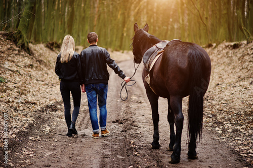 Back of young stylish couple in love walking with horse at autumn forest.