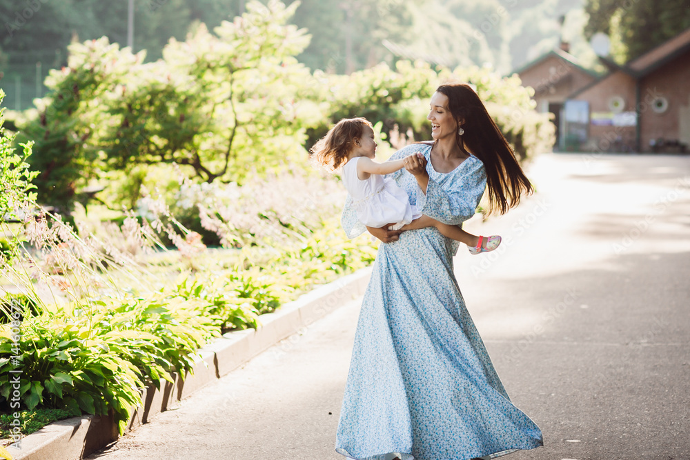 Woman in long blue dress whirls with her little daughter on pavement path