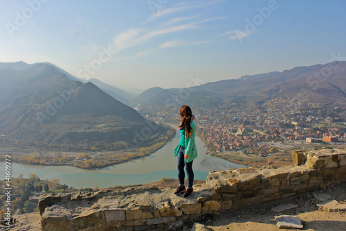 Traveler positive girl standing on the stone wall. Territory Jvari Monastery. City Mtskheta, rivers Aragvi and Kura, and stunning mountains at the background. Freedom concept. photo